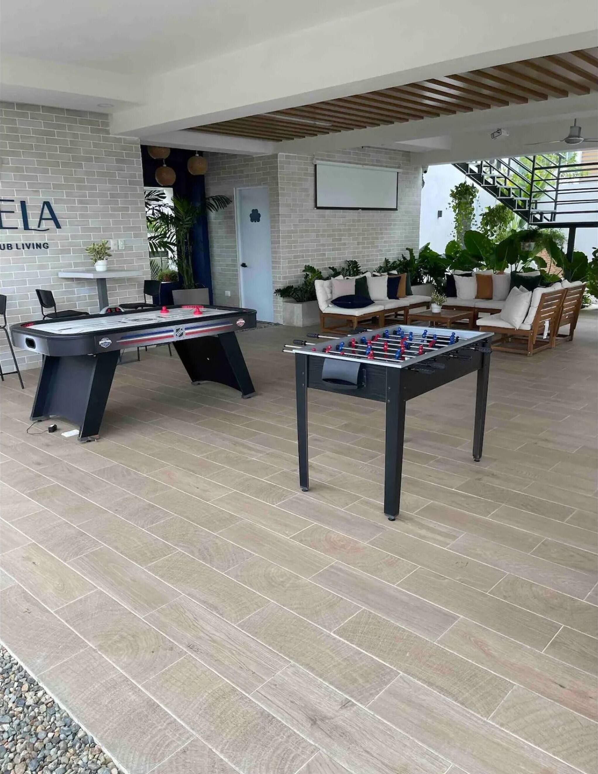 Una mesa de billar, una mesa de futbolín y un sofá en un área de descanso con pared de ladrillo y plantas.