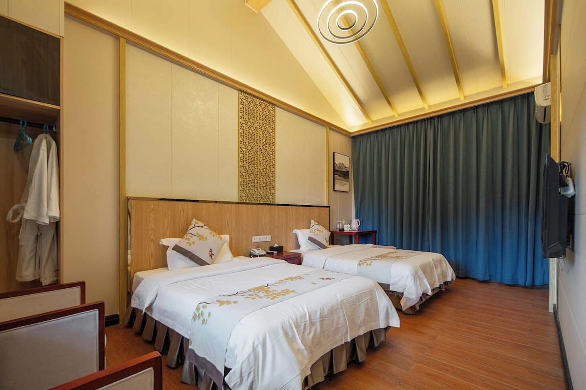 Une chambre d’hôtel avec deux lits, une tête de lit en bois, un panneau décoratif au mur, un luminaire au plafond, une table de chevet et une fenêtre avec des rideaux.