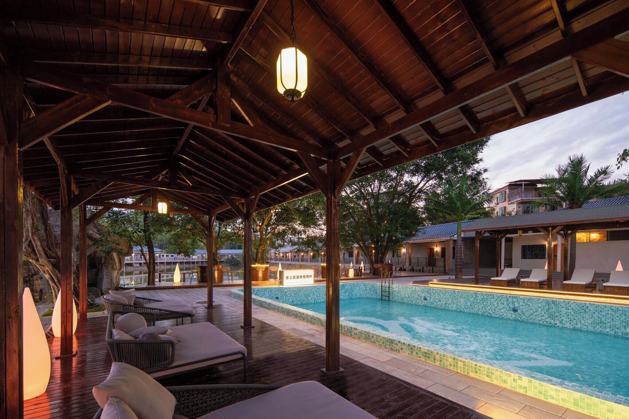 Un área de piscina techada con sillones de descanso, una terraza de madera y vistas a un edificio al fondo.