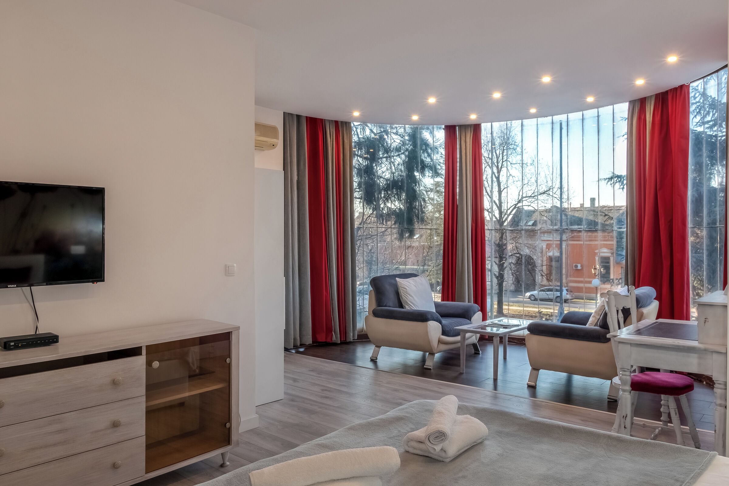Un salon moderne doté d’un téléviseur à écran plat, d’un meuble en bois, de deux fauteuils, d’une table basse et d’un coin repas avec une table blanche et des rideaux rouges.