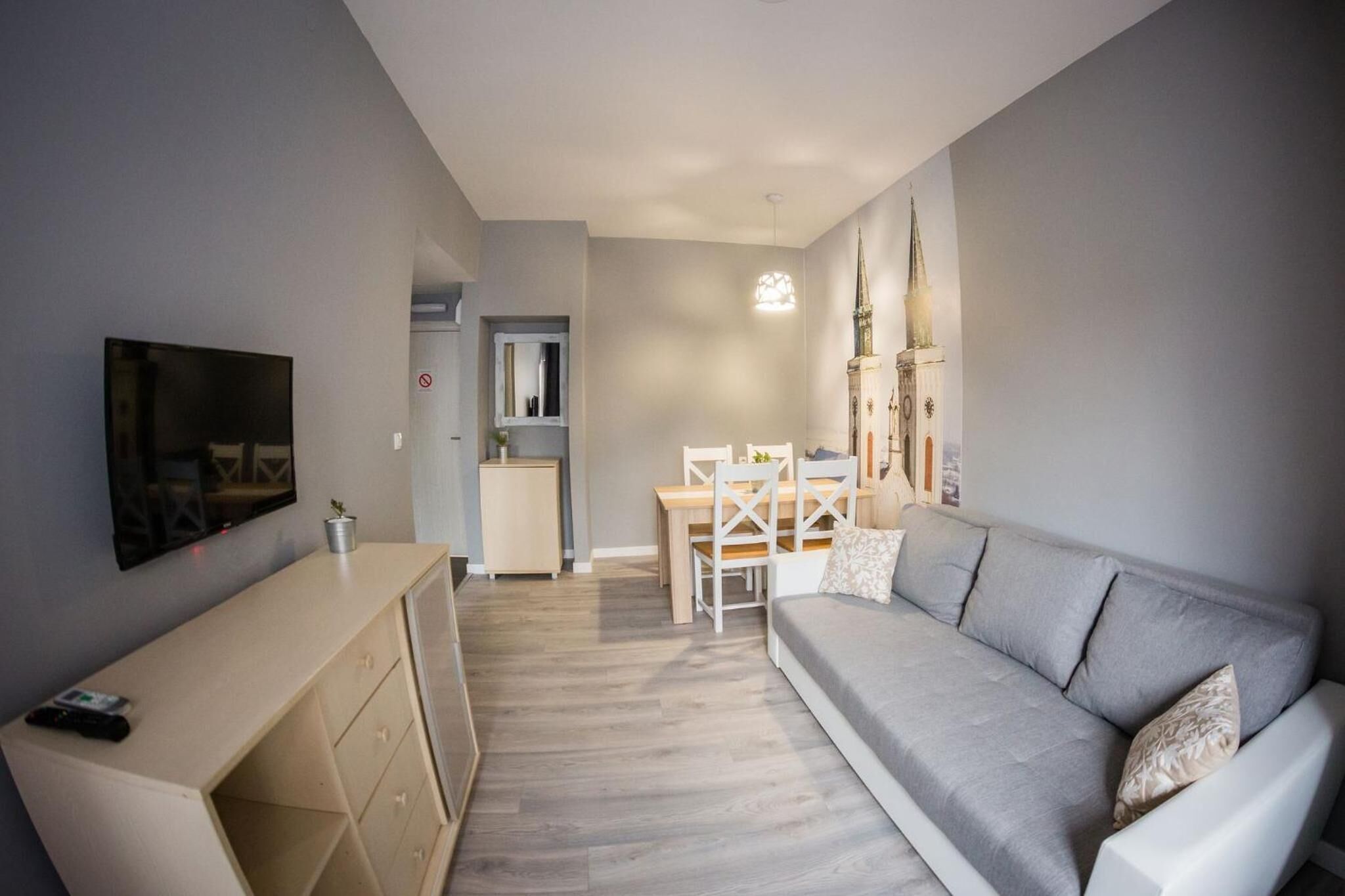 Een woonkamer met een grijze bank, een televisie, een houten eettafel en een klein keukentje.
