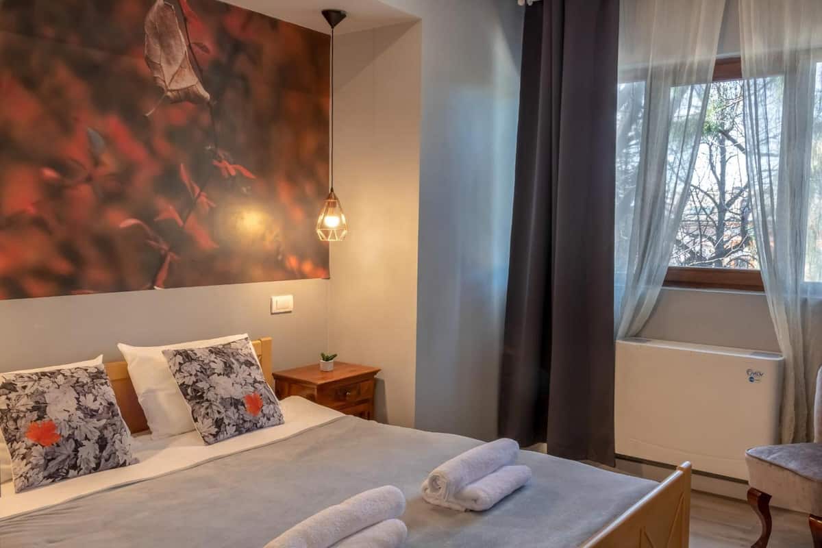 A hotel room with a bed, pillows, a nightstand, a lamp, a window with curtains, and an air conditioning unit.