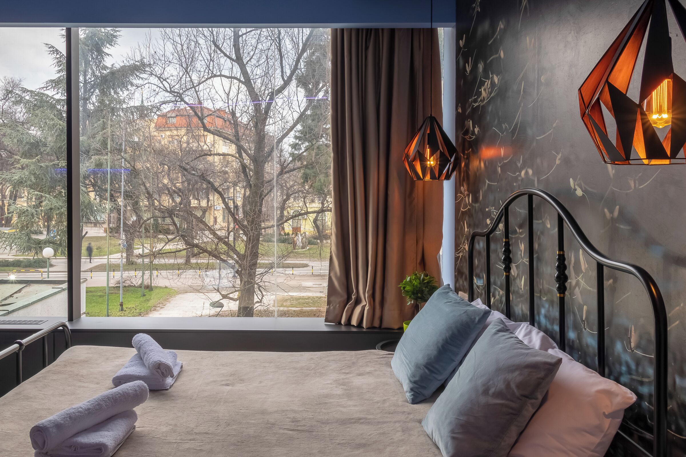 A bedroom with a large window, a bed, a nightstand, and a hanging light fixture.