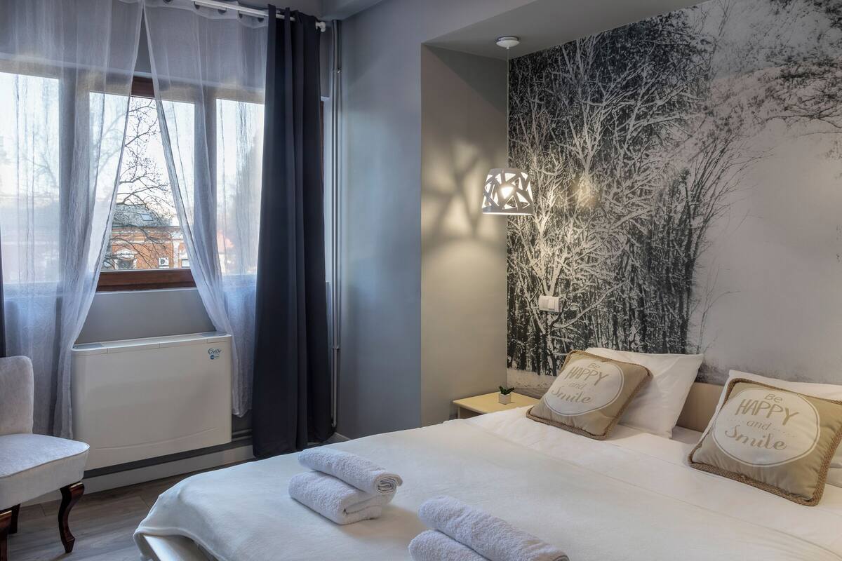 A hotel room with a bed, a chair, a radiator, a wall with a tree mural, and a nightstand with a lamp.
