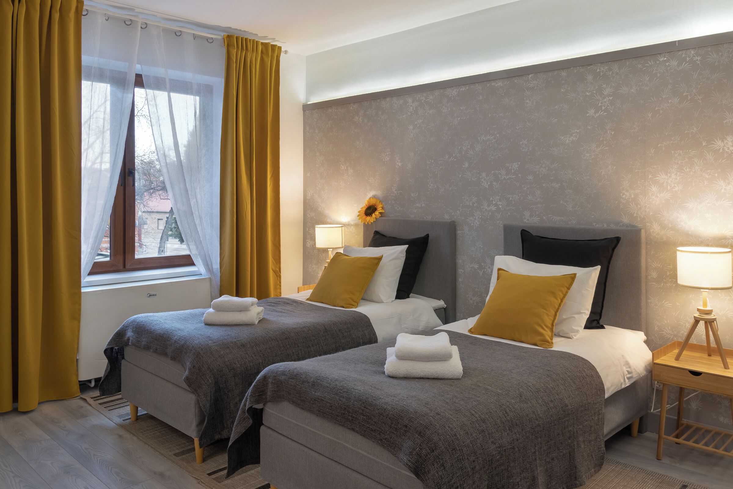 A hotel room with two beds, a window with yellow curtains, a bedside table with a lamp, and a wall with a floral pattern.