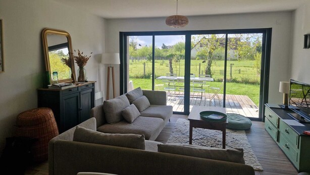 Una sala de estar con sofá, mesa, lámpara, espejo y vista a un jardín a través de puertas de cristal amplias.
