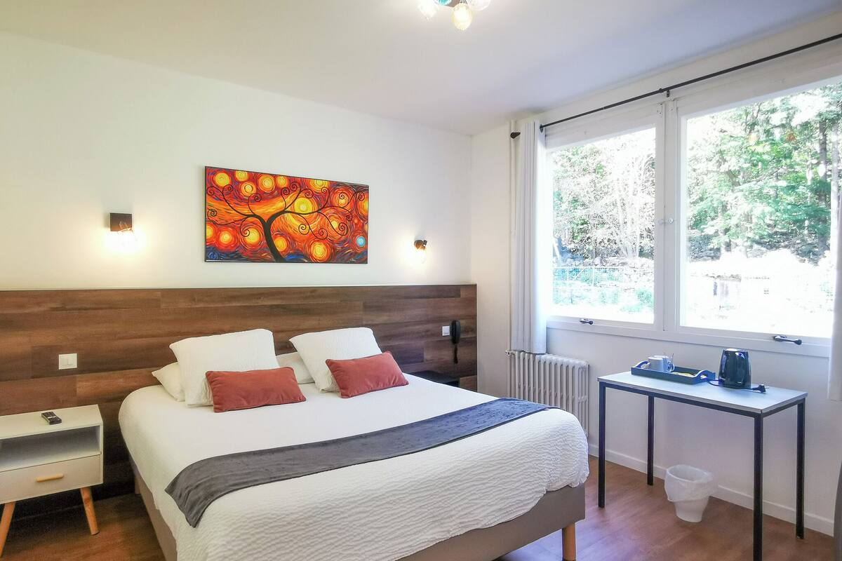 Een slaapkamer met een bed, nachtkastje, radiator en een raam met uitzicht op bomen.