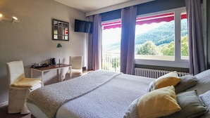 A hotel room with a large bed, a desk with a chair, a TV mounted on the wall, and a window with a view of the outdoors. - Hôtel Restaurant le Tanargue (Valgorge)