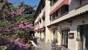 An outdoor cafe area with tables and chairs, a staircase leading to an upper floor, and a tree with purple flowers. - Hôtel Restaurant le Tanargue (Valgorge)