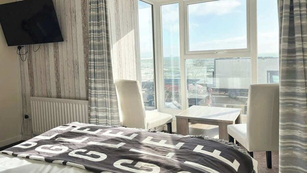 Ein Hotelzimmer mit einem Bett, einem an der Wand befestigten Fernseher, einem kleinen Tisch, zwei Stühlen und einem großen Fenster mit Blick auf die Stadt.