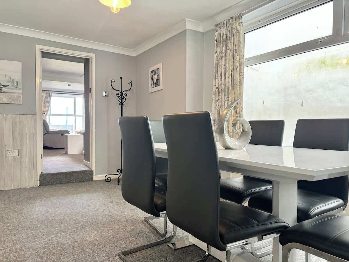 A dining area with a white table and black chairs, a window with curtains, and a door leading to another room.