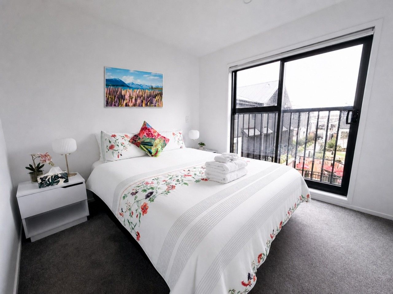 A bedroom with a large window, a bed with floral bedding, a nightstand with a lamp, and a painting on the wall.