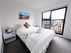 Ein Schlafzimmer mit großem Fenster, einem Bett mit Blumenmuster, einem Nachttisch mit Lampe und einem Gemälde an der Wand.