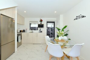 A modern kitchen with a built-in oven, a dining area with a glass table and white chairs, and a view of the outdoors through a door. - Spacious 2BR Townhouse with Yard Wilmer Street (Christchurch)