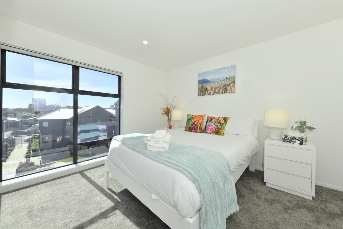 A modern bedroom with a large window, a bed with white linens, colorful pillows, a bedside table with a lamp, and a painting on the wall.