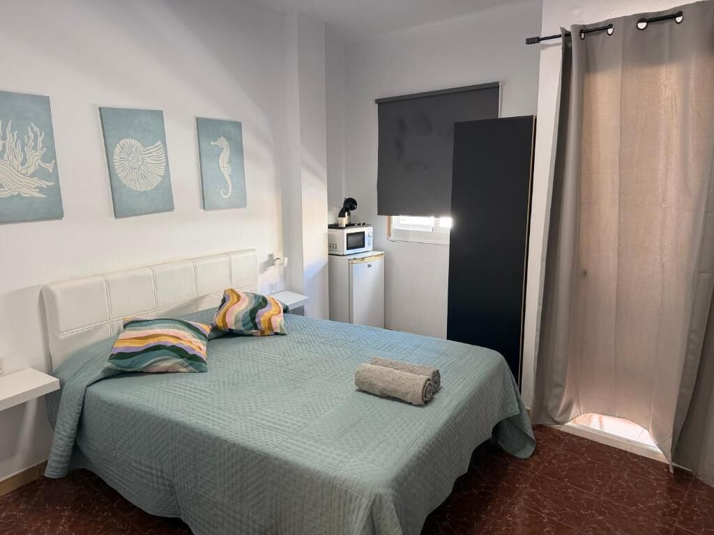 A bedroom with a bed, a bedside table, a microwave, and a window with a curtain.