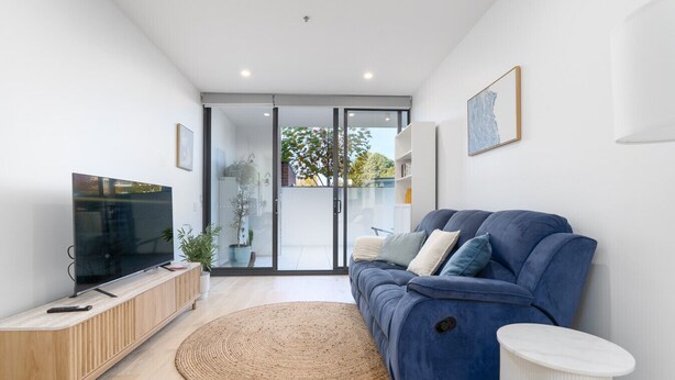 A modern living room with a flat-screen TV, a blue sofa, a wooden cabinet, and a large window. - Griffith Apartment | Prime Location+Courtyard+Gym (Griffith)