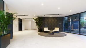 A modern lobby with a reception desk, two seating areas, a large potted plant, and a glass wall with a view. - Griffith Apartment | Prime Location+Courtyard+Gym (Griffith)