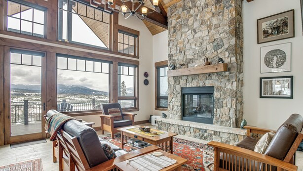 Un salón amplio con chimenea de piedra, muebles de madera, ventanas grandes y vistas a la montaña.