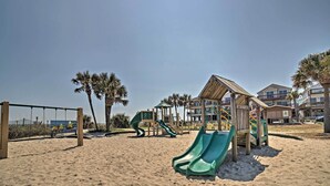 Ein Spielplatz am Strand mit Rutschen und einer Holzkonstruktion, umgeben von Sand und Palmen.