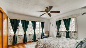Un dormitorio con una cama, un ventilador de techo, una ventana con cortinas, una silla y una alfombra.