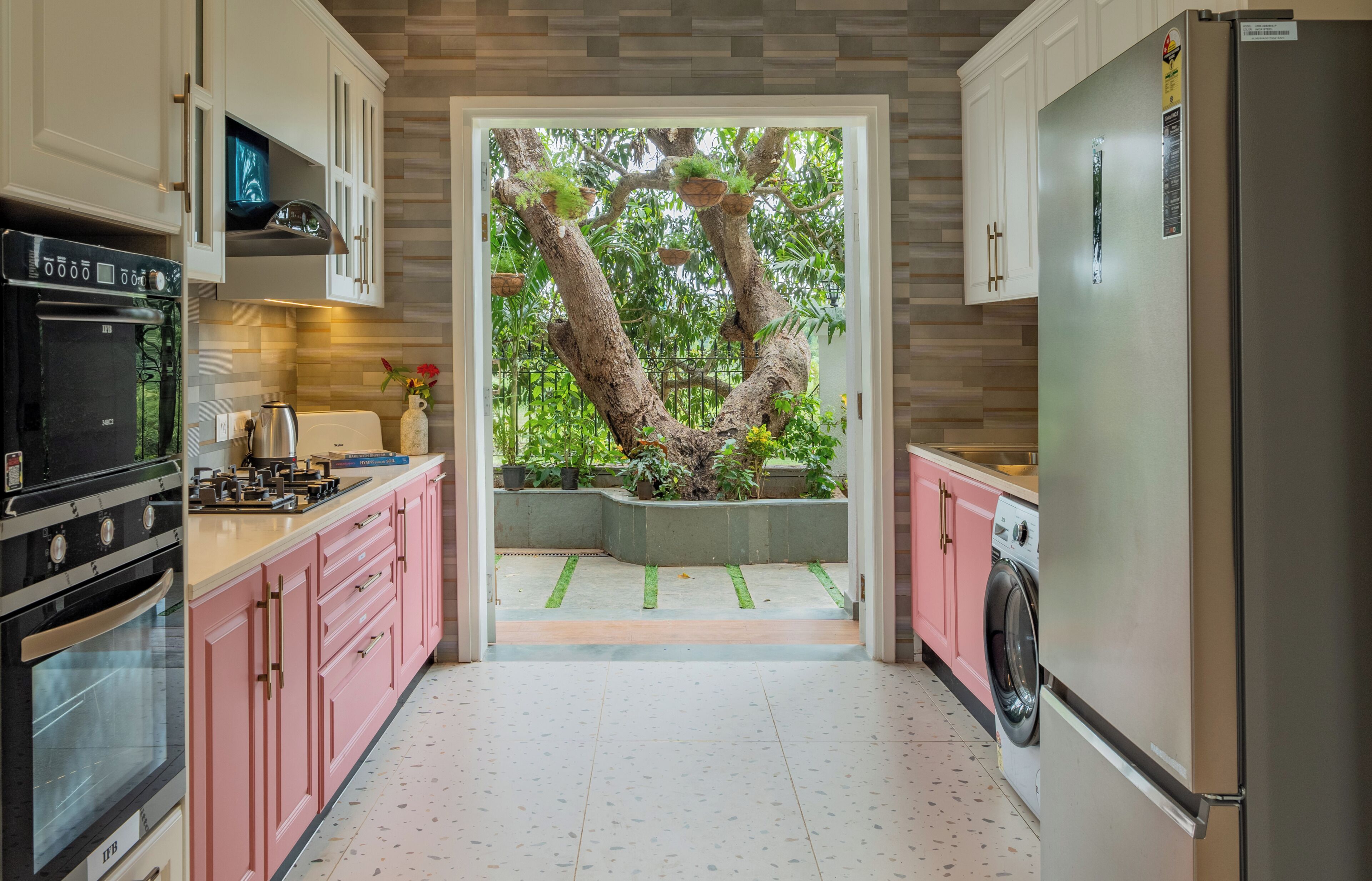 A kitchen with white cabinets, a stainless steel refrigerator, a washing machine, and a tree planter with plants.