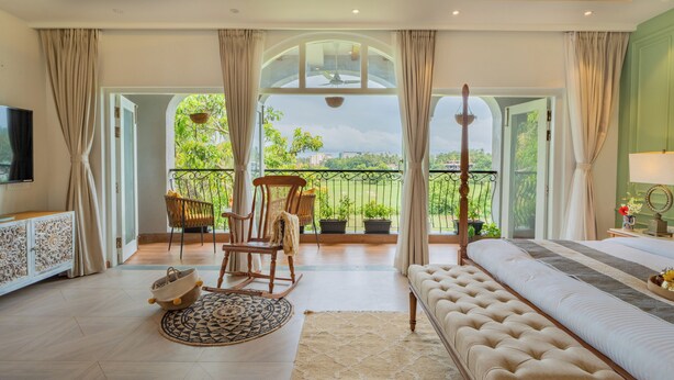 Un dormitorio amplio con una cama grande, televisión, balcón con sillas y plantas en macetas, y vistas al verdor.