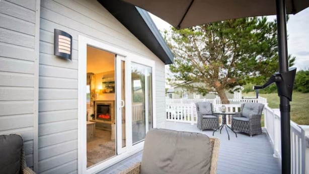 A covered deck with wicker furniture, a sliding glass door leading to a room with a fireplace, and a white fence in the background. - Meadowbank Lodge with ceiling surround system nr beach pet friendly Shorefields (Milford on Sea)