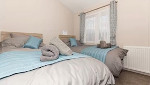 A hotel room with two beds, a window with curtains, a radiator, and a wall-mounted light switch. - Meadowbank Lodge with ceiling surround system nr beach pet friendly Shorefields (Milford on Sea)