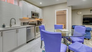 Une cuisine moderne avec des meubles blancs, des appareils en acier inoxydable et une table à manger en verre, assortie de chaises violettes.