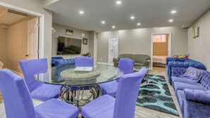 Un salon moderne avec une table à manger au plateau en verre, entourée de chaises violettes, d’un canapé bleu et d’un tapis à motifs léopard.