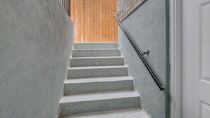 Un escalier avec une rampe en bois en arrière-plan, une main courante en métal et une porte blanche sur la droite.