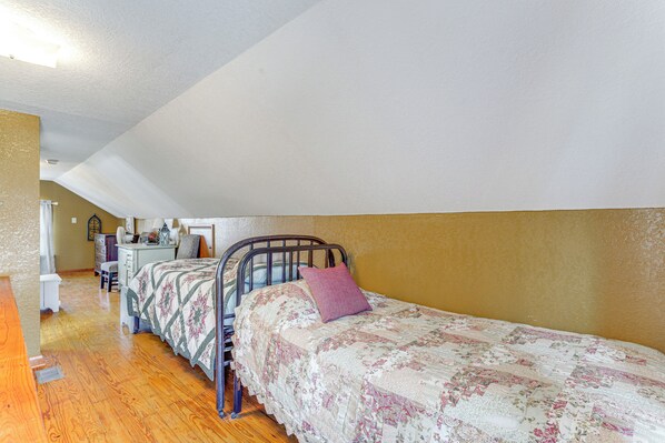 A bedroom with a sloped ceiling, a bed with a floral quilt, a chair, and a desk with a lamp. - Half-Mi to Boat Ramp! Gasconade Country Cottage (Gasconade)