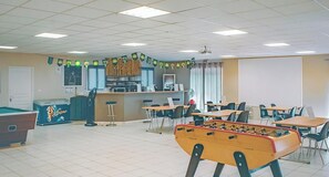 A room with a pool table, a foosball table, and a bar area with a refrigerator and stools.