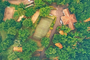 Vista aérea de una zona residencial con cancha de tenis, una casa grande de techo peculiar y vegetación circundante.