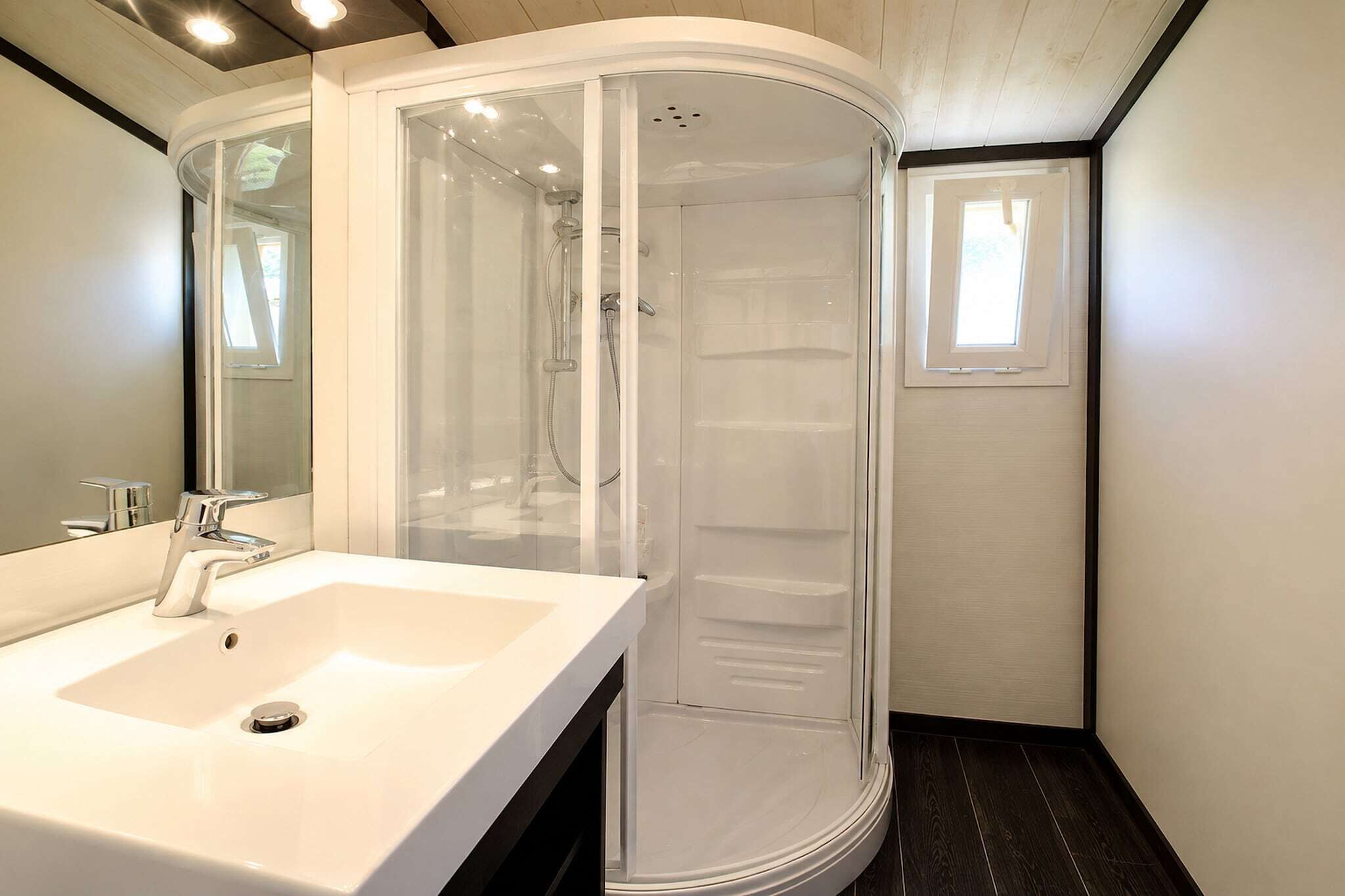 Un baño moderno con un lavamanos blanco, ducha con mampara de vidrio y una pequeña ventana.