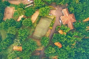 Vista aérea de una zona residencial con cancha de tenis, una casa grande de techo peculiar y vegetación circundante.
