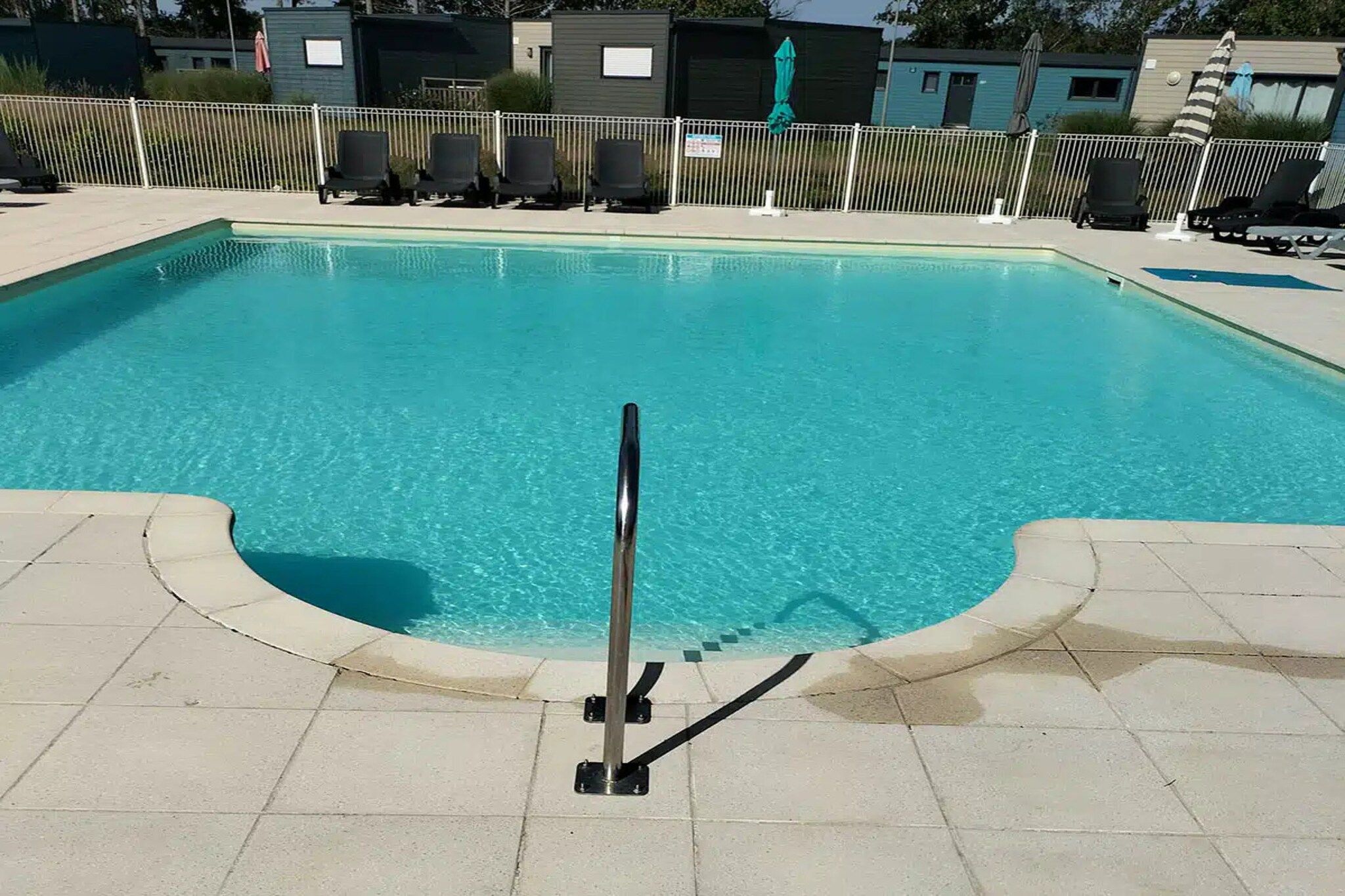 Piscina com escada de metal dando acesso à água, cercada por uma grade branca e cadeiras de sol.