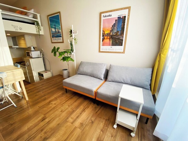 Ein gemütlicher Wohnraum mit einer Couch, einem kleinen Tisch, einer Mikrowelle, einem Kühlschrank, einer Topfpflanze und zwei gerahmten Bildern an der Wand.