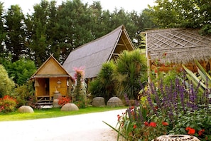A traditional wooden house with a thatched roof, surrounded by lush greenery and flowering plants.