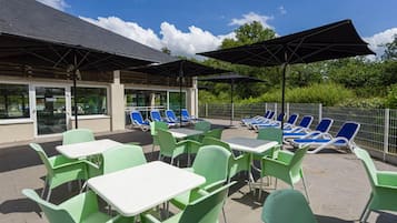 An outdoor patio with green chairs and tables, umbrellas, and a pool area with blue lounge chairs.