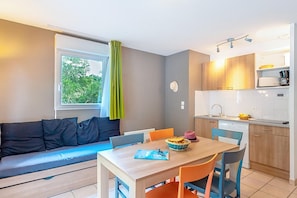 Ett kompakt bostadsutrymme med en soffa, ett matbord, stolar, ett litet köksområde och ett fönster med gardiner.