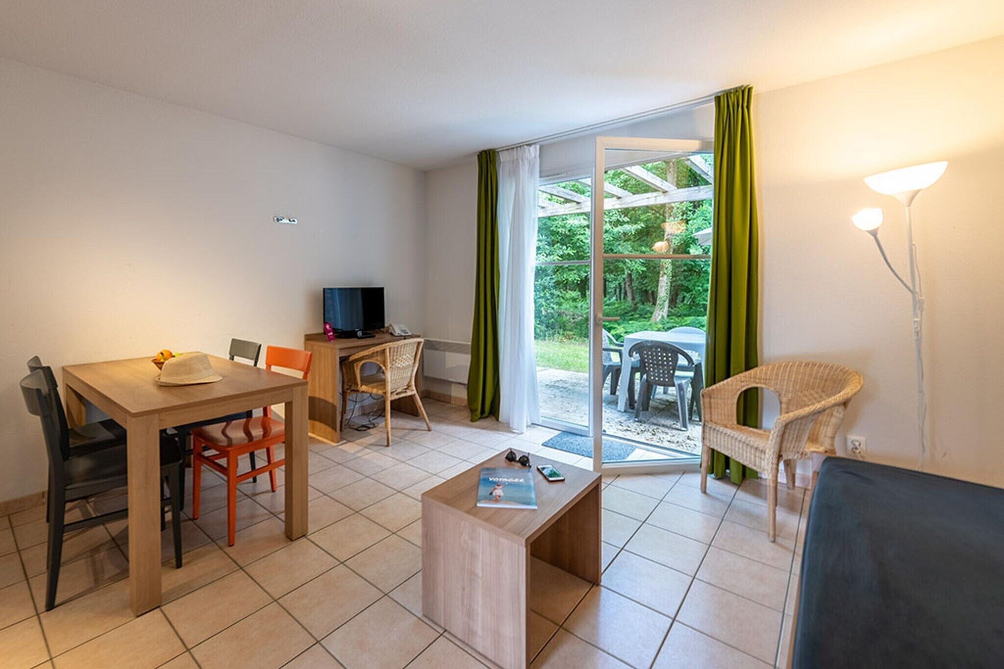Ein Zimmer mit Esstisch, Stühlen, Fernseher, Sofa und Blick ins Grüne durch eine Glastür.