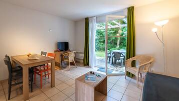 Ein Zimmer mit Esstisch, Stühlen, Fernseher, Sofa und Blick ins Grüne durch eine Glastür.