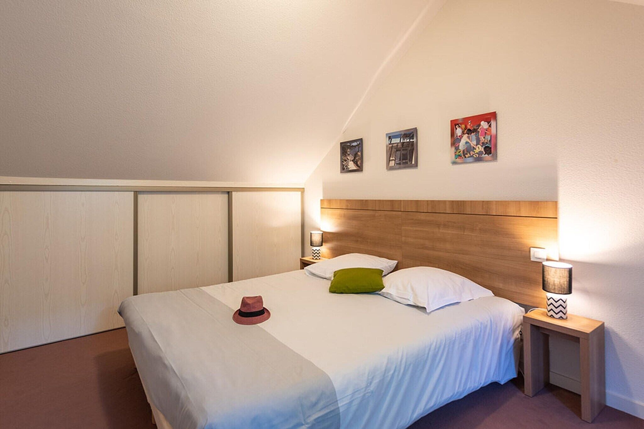 Ein ordentlich bezogenes Bett mit weißer Bettwäsche, einem grünen Kissen und einem roten Hut. Zwei Nachttische mit Lampen stehen daneben, und der Raum hat eine geneigte Decke mit Holzverkleidung.