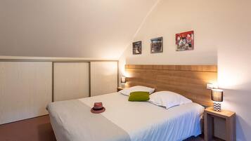 Ein ordentlich bezogenes Bett mit weißer Bettwäsche, einem grünen Kissen und einem roten Hut. Zwei Nachttische mit Lampen stehen daneben, und der Raum hat eine geneigte Decke mit Holzverkleidung.
