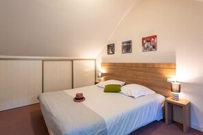 A neatly made bed with white bedding, a green pillow, and a red hat on the bed. There are two bedside tables with lamps, and the room has a sloped ceiling with wooden paneling.