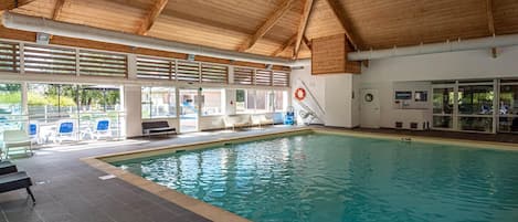An indoor swimming pool with clear water, surrounded by lounge chairs and a covered roof structure.