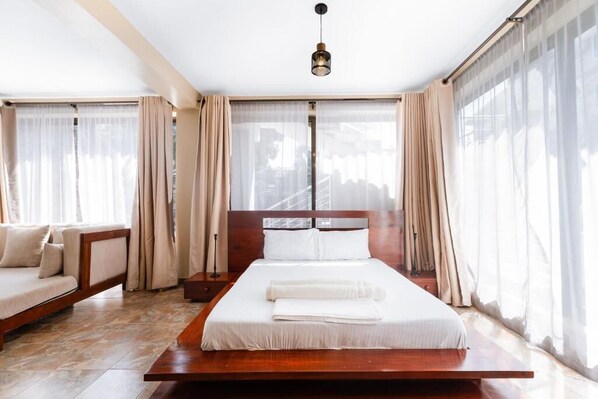 Habitación de hotel moderna con una cama grande, un sofá y vistas a un edificio a través de cortinas translúcidas.