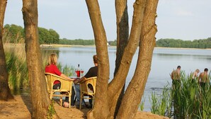 Ein Seeufer mit Tisch und Stühlen unter Bäumen, mit Blick auf einen stillen See.